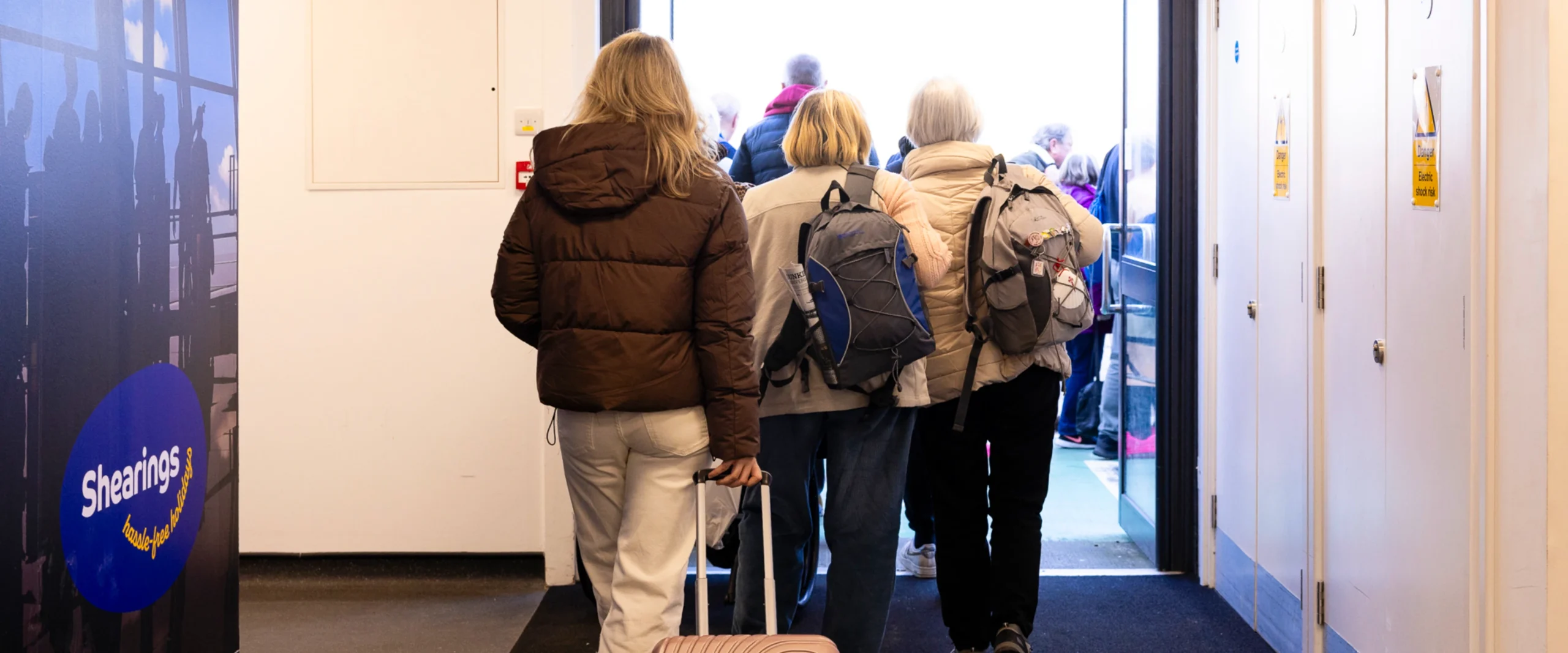 A group of passengers walk through a bright corridor towards a waiting coach outside. The traveller at the back of the group pulls a small suitcase while others carry backpacks. A large Shearings Holidays logo on the left wall is visible, suggesting an organised group tour. The moment captures the smooth transition from indoor facilities to coach boarding.