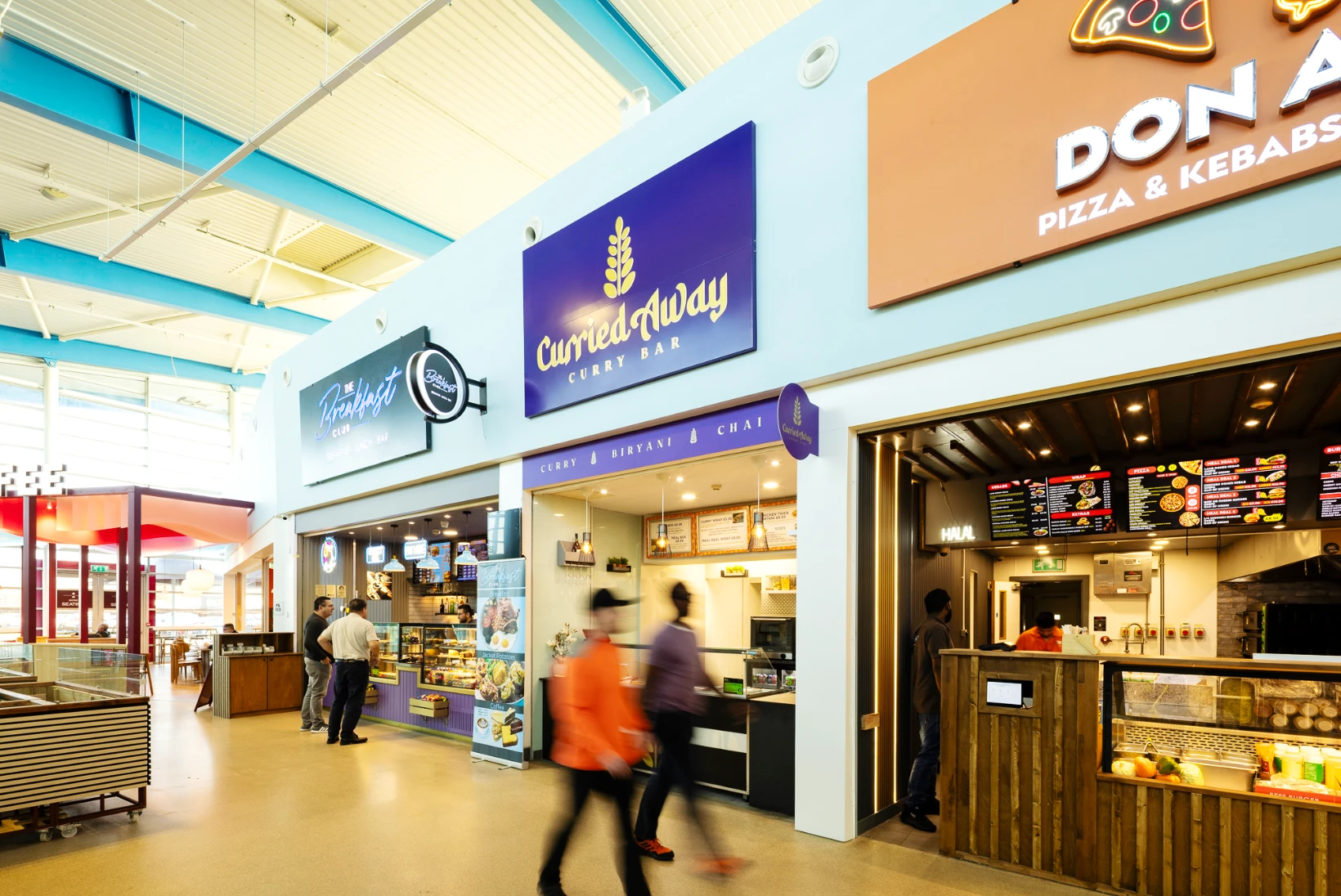 An interior view of the Folkestone Services food court shows three visible diverse eateries: The Breakfast Club, Curried Away Curry Bar, and Don Air Pizza & Kebabs. The space is bright with natural light from the high glass windows and a sky-blue ceiling. A few customers browse food displays while walking past, creating a sense of motion and activity.