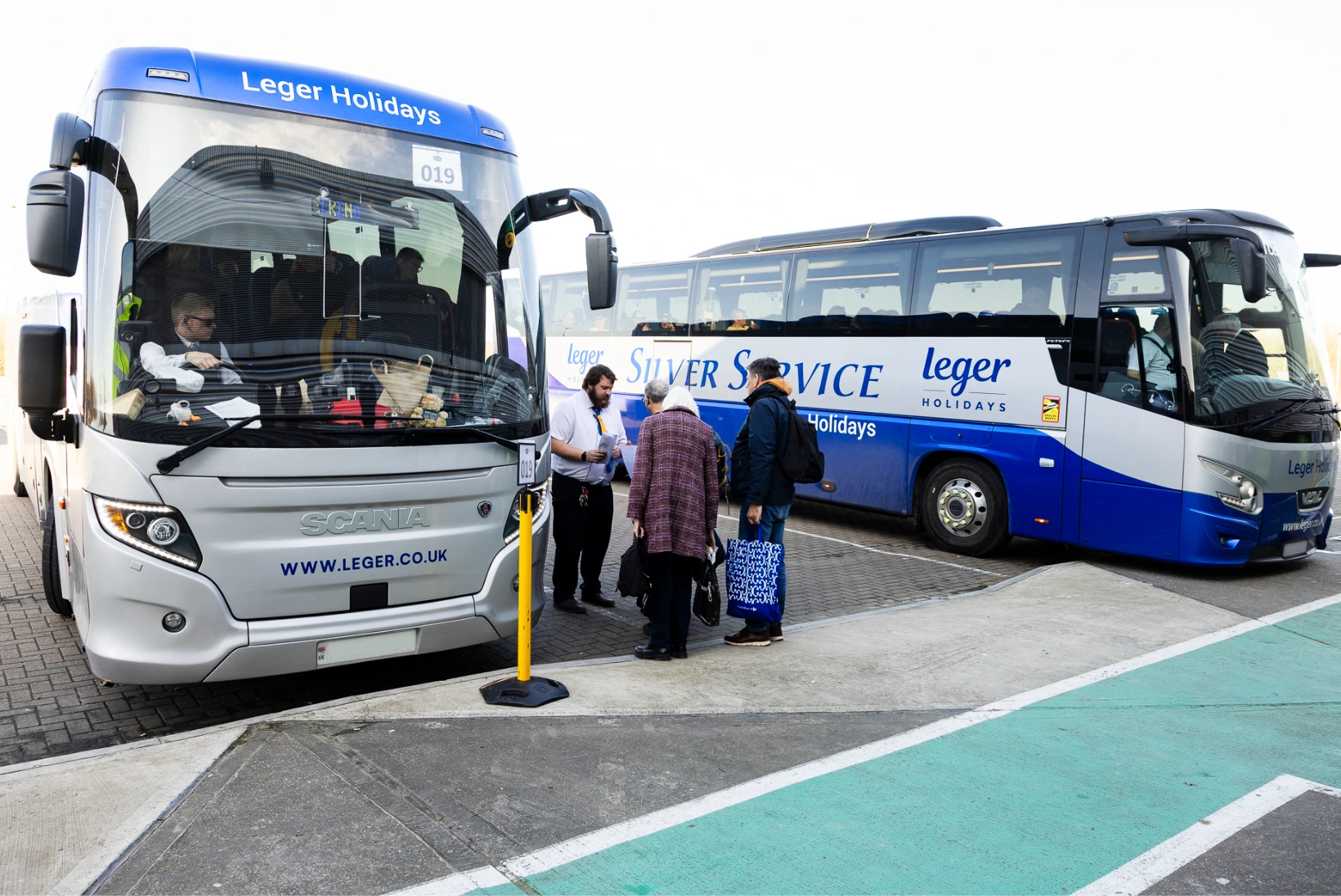 Two Leger Holidays coaches are parked side by side in a designated coach parking area. One driver is seated in the cab while a staff member checks passenger paperwork next to the coach. Three passengers stand by with bags, ready to board. The area is clean and well-marked, providing a clean, organised environment for safe boarding and disembarkation.