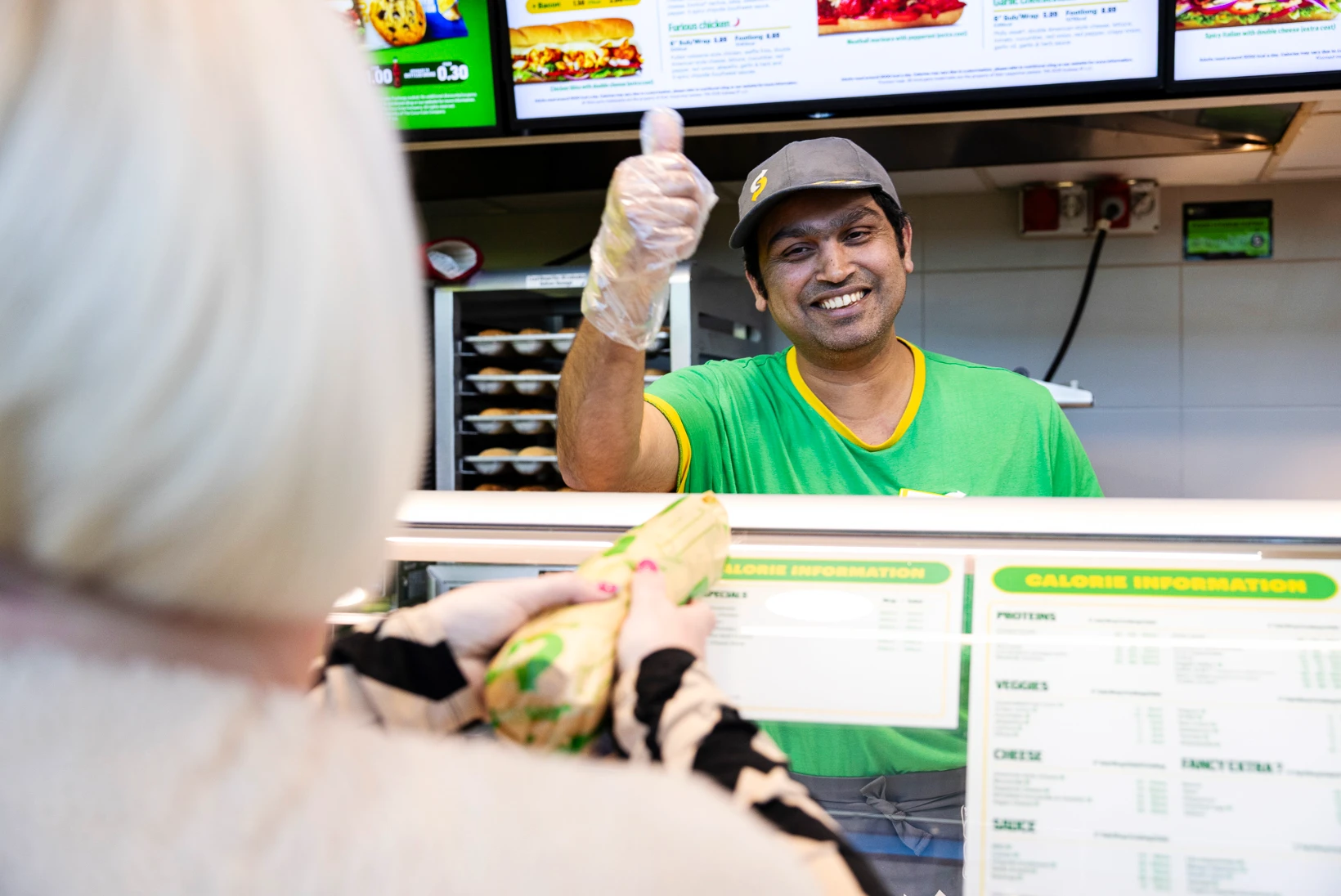 A cheerful Subway staff member in a green uniform and cap gives a thumbs-up from behind the counter while smiling at a customer. The customer, seen from behind, receives a freshly wrapped sub. Digital menu boards with meal options and calorie information are visible overhead. The image conveys friendly, fast service and a welcoming food experience for travellers.