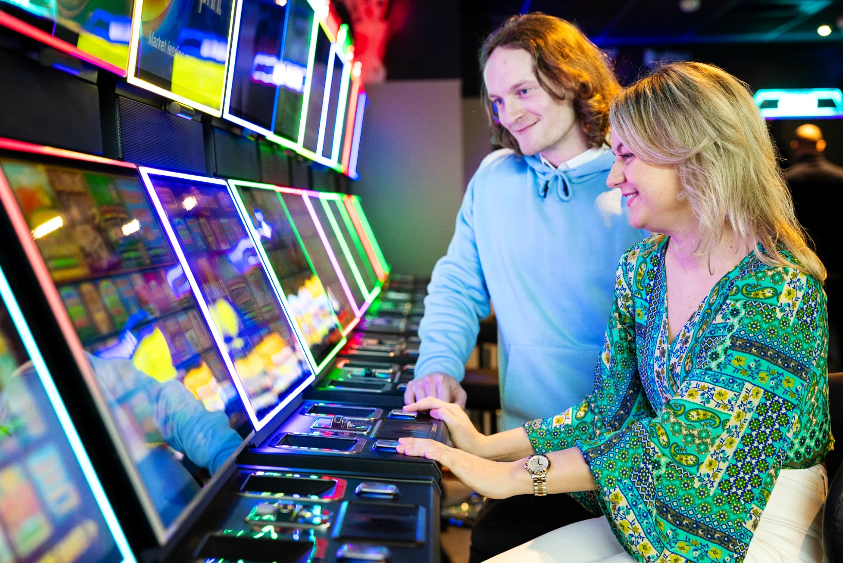 A smiling woman and man enjoy a row of colourful, illuminated slot machines in a gaming lounge. The bright neon lights reflect off the screens, creating a lively and energetic atmosphere ideal for travellers looking to unwind with some entertainment at Folkestone Services.