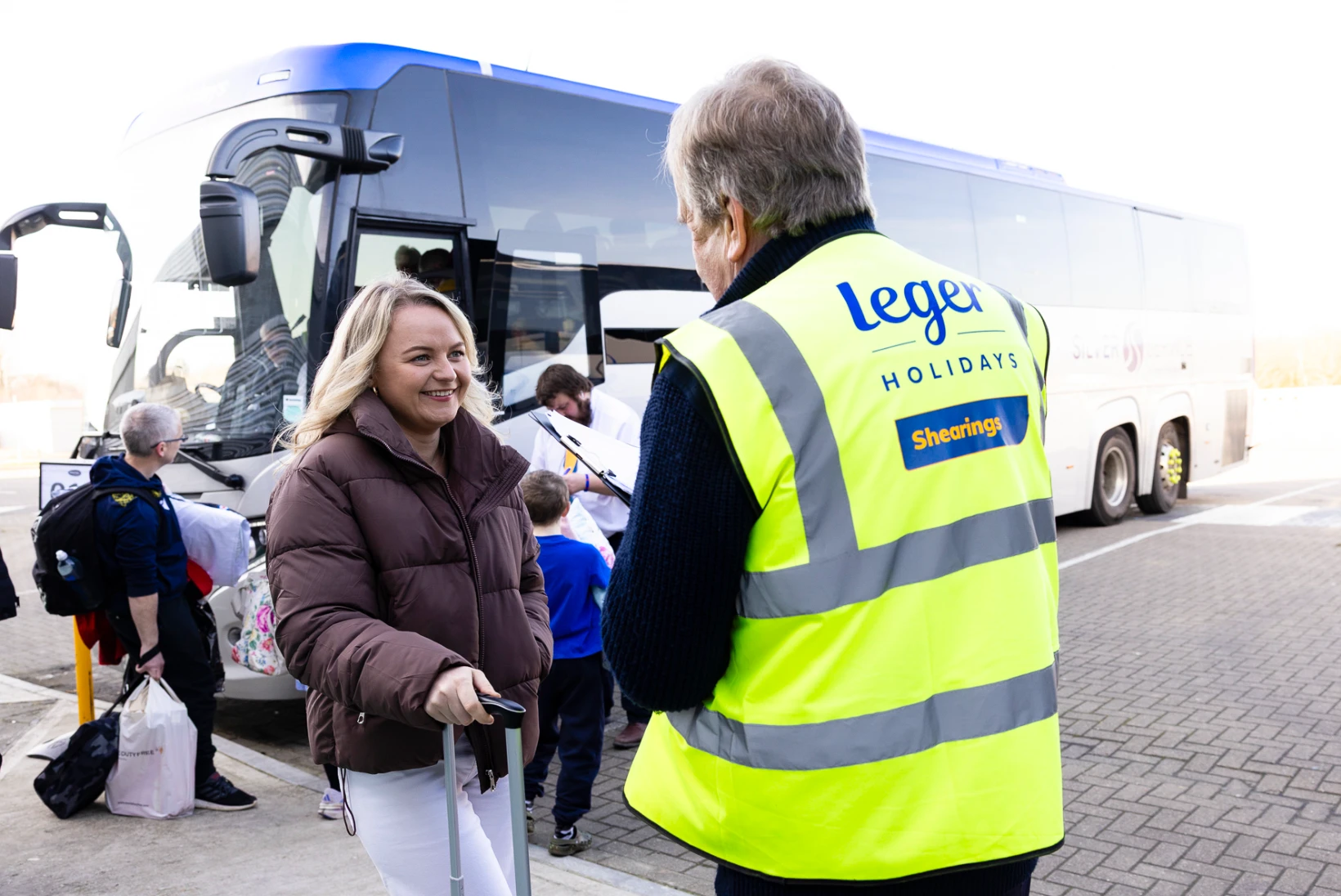 A smiling woman pulls a suitcase while speaking with a Leger Holidays staff member wearing a bright yellow high-vis vest. In the background, passengers and children gather around two parked coaches, preparing for departure. The scene captures the organised and friendly atmosphere of the designated coach parking area at Folkestone Services.
