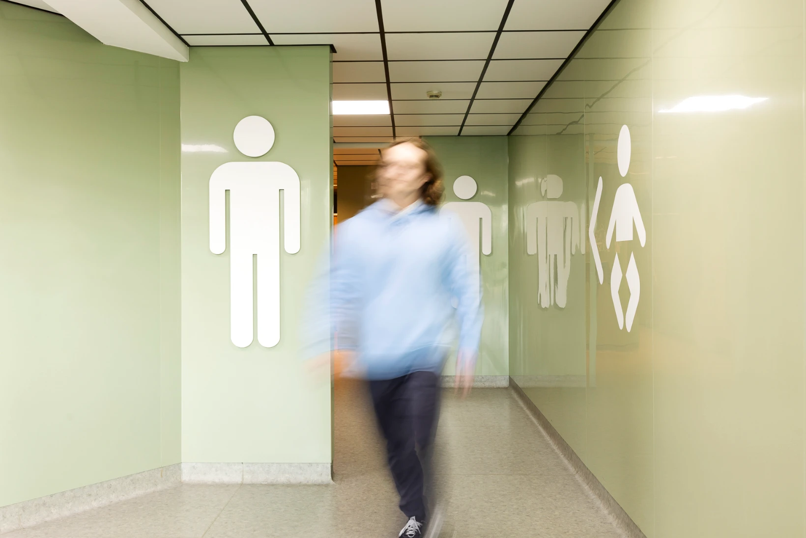 A blurred male figure walks through a well-lit corridor with pastel green walls at Folkestone Services. Large, white pictograms for male and female restrooms are visible on both sides of the hallway, guiding visitors toward the clean and modern toilet facilities.