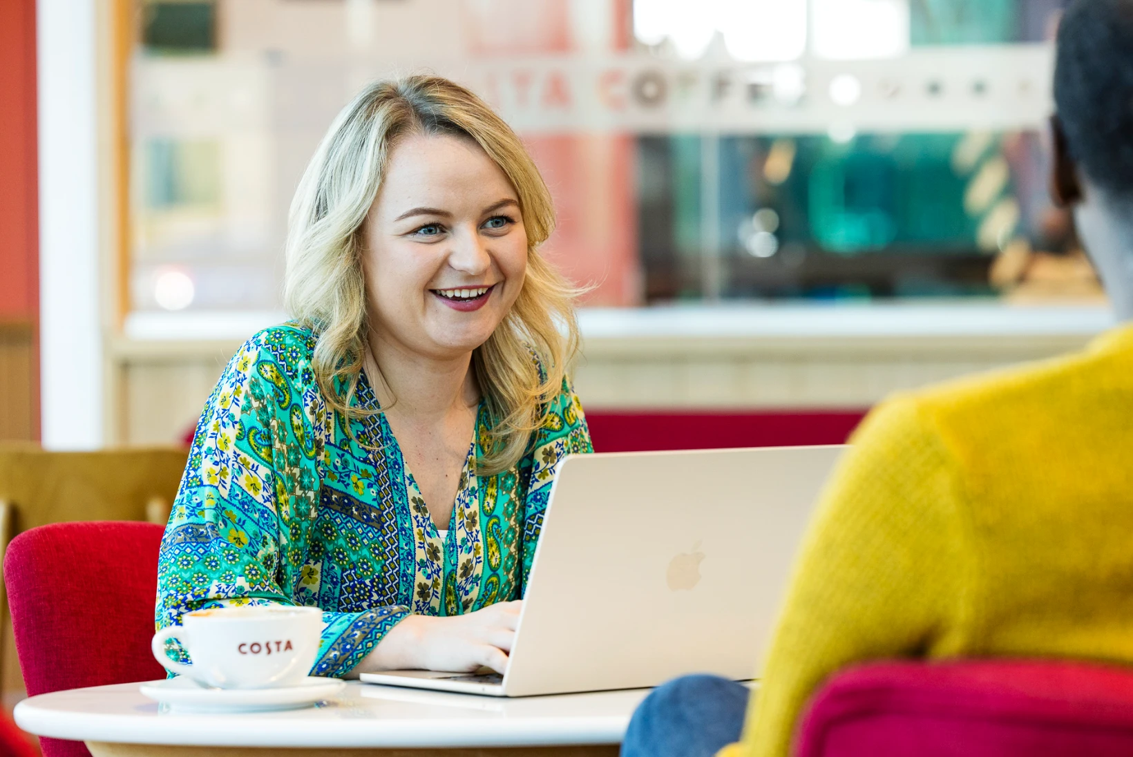 A smiling woman sits at a round table in a Costa Coffee lounge, working on a laptop with a coffee cup in front of her. The relaxed café setting, with soft red seating and ambient lighting, highlights the free high-speed Wi-Fi available at Folkestone Services.